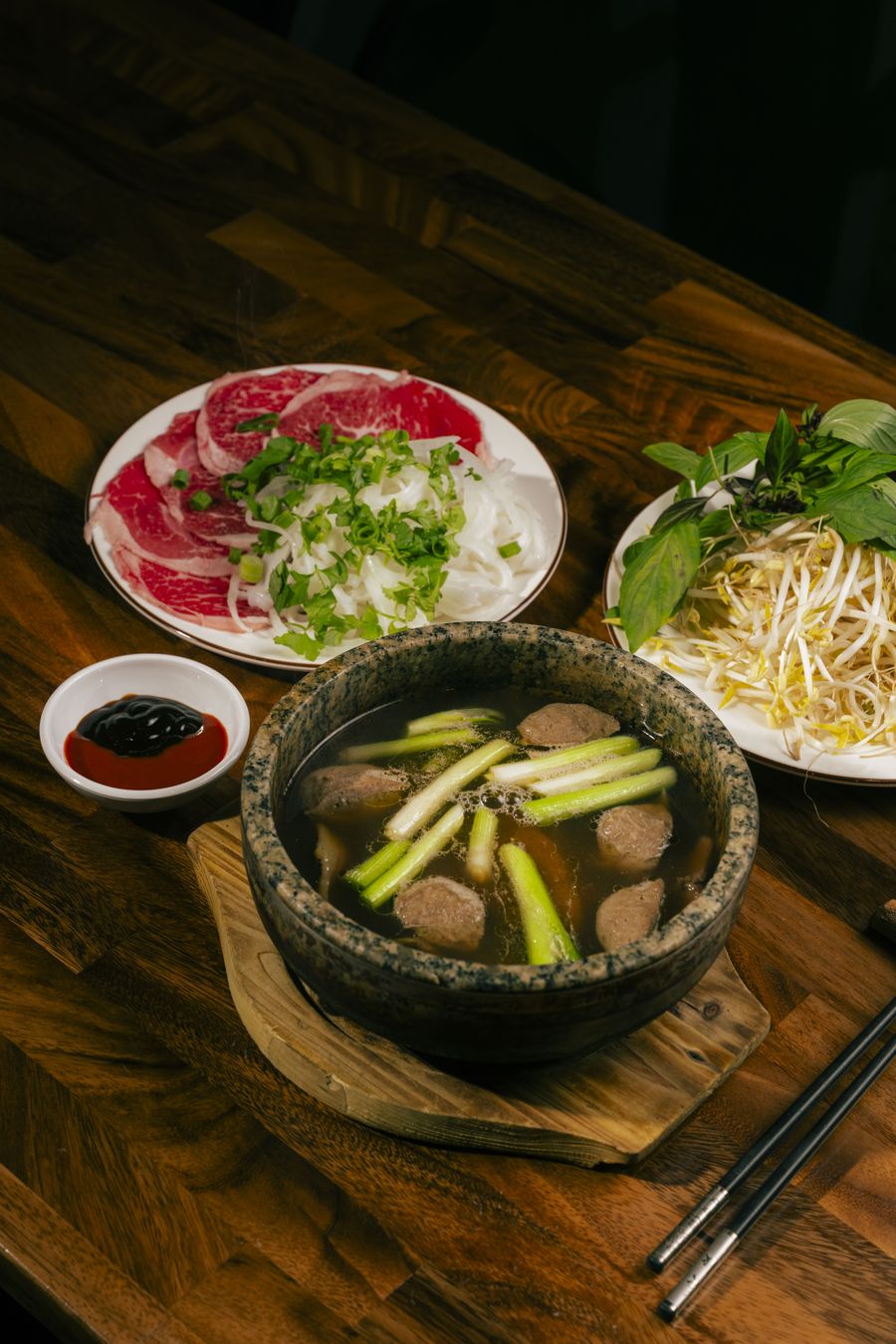 Stone bowl of 18-hour bone broth with beef balls and spring onion, beside a plate of rare beef and herbs